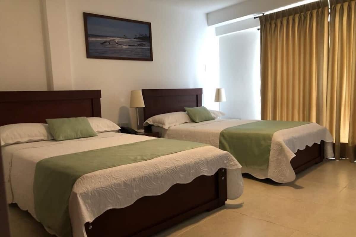A hotel room with two beds, a painting on the wall, a window with curtains, and a lamp on the nightstand.