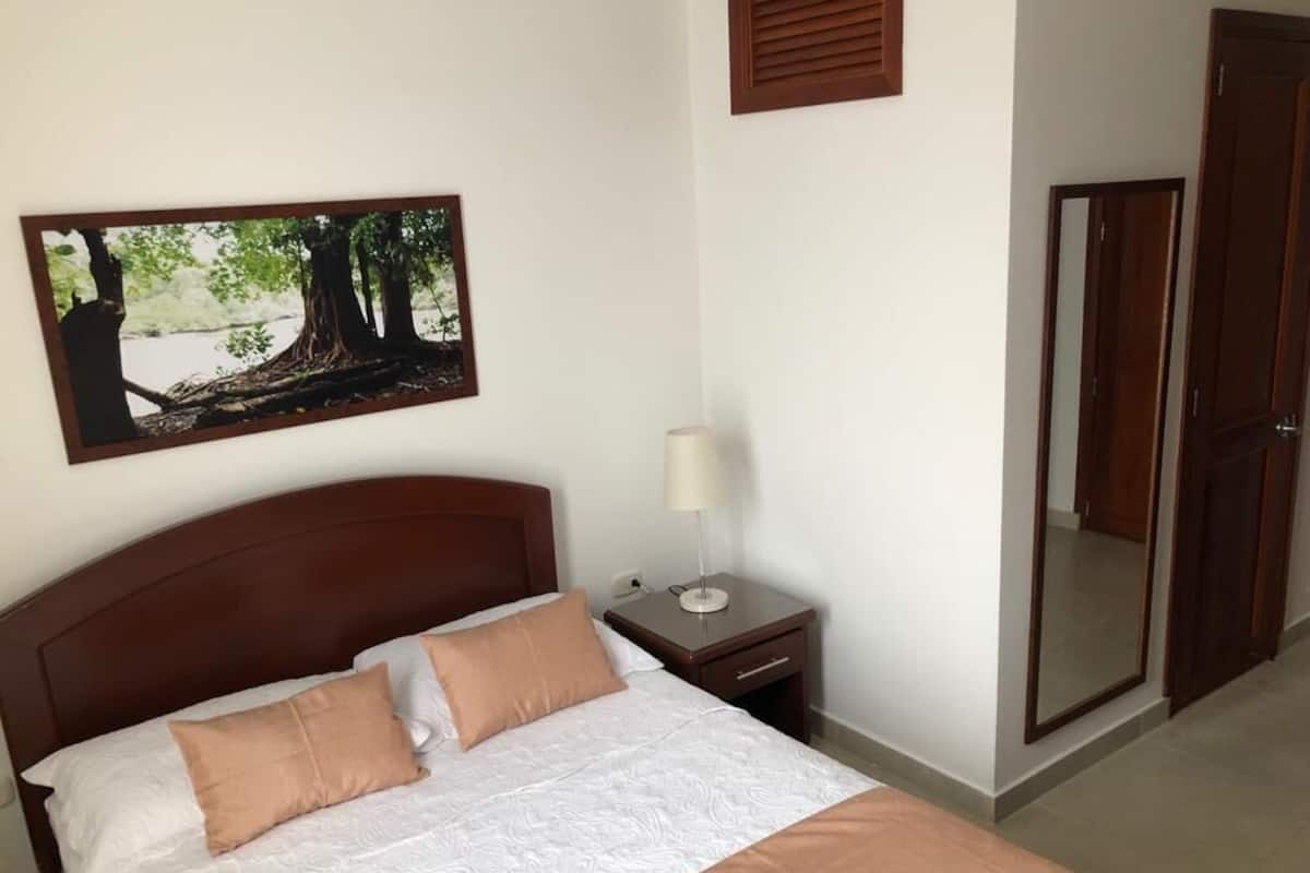 A neatly made bed with a headboard, a bedside table with a lamp, a framed picture on the wall, and a door with a mirror.
