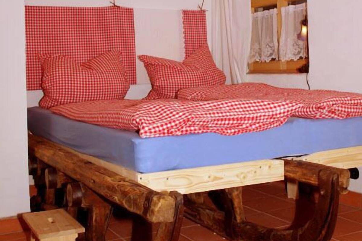 A rustic wooden bed with a checkered red and white bedding, a small wooden stool, and a window with white curtains.