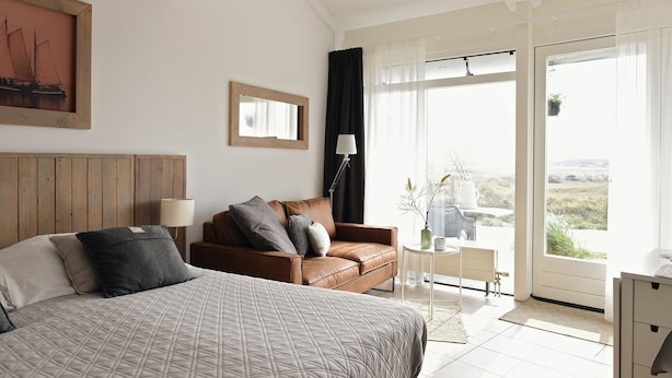 A bedroom with a bed, a sofa, a small table, a lamp, and a window with a view. - De Duynsuites (Callantsoog)