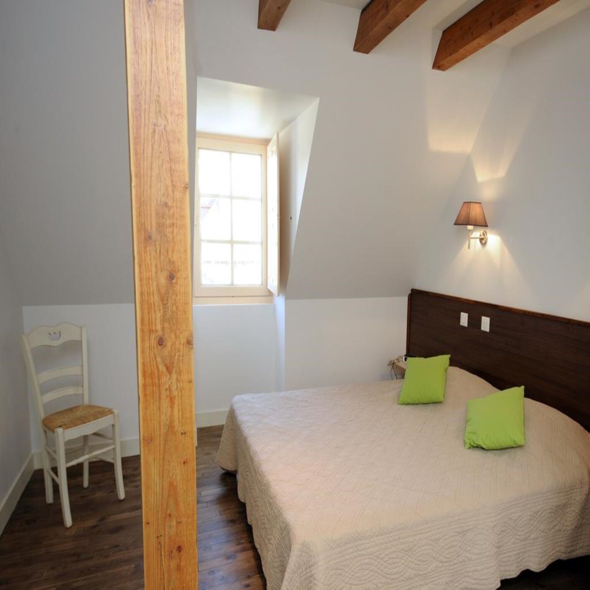 Ein Schlafzimmer mit einem hölzernen Kopfteil, einem Bett mit zwei grünen Kissen, einem weißen Stuhl und einem Fenster mit Tageslicht.
