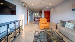 A spacious living room with a sofa, a coffee table, and a TV mounted on the wall. There is a ceiling fan and a kitchen visible in the background. - Coastal-Inspired Condo w/ Infinity Pool & Oceanview Terrace (Playa Flamingo)