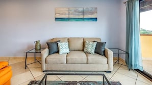 A living room with a beige sofa, patterned cushions, a glass coffee table, and a painting of a seascape. - Coastal-Inspired Condo w/ Infinity Pool & Oceanview Terrace (Playa Flamingo)