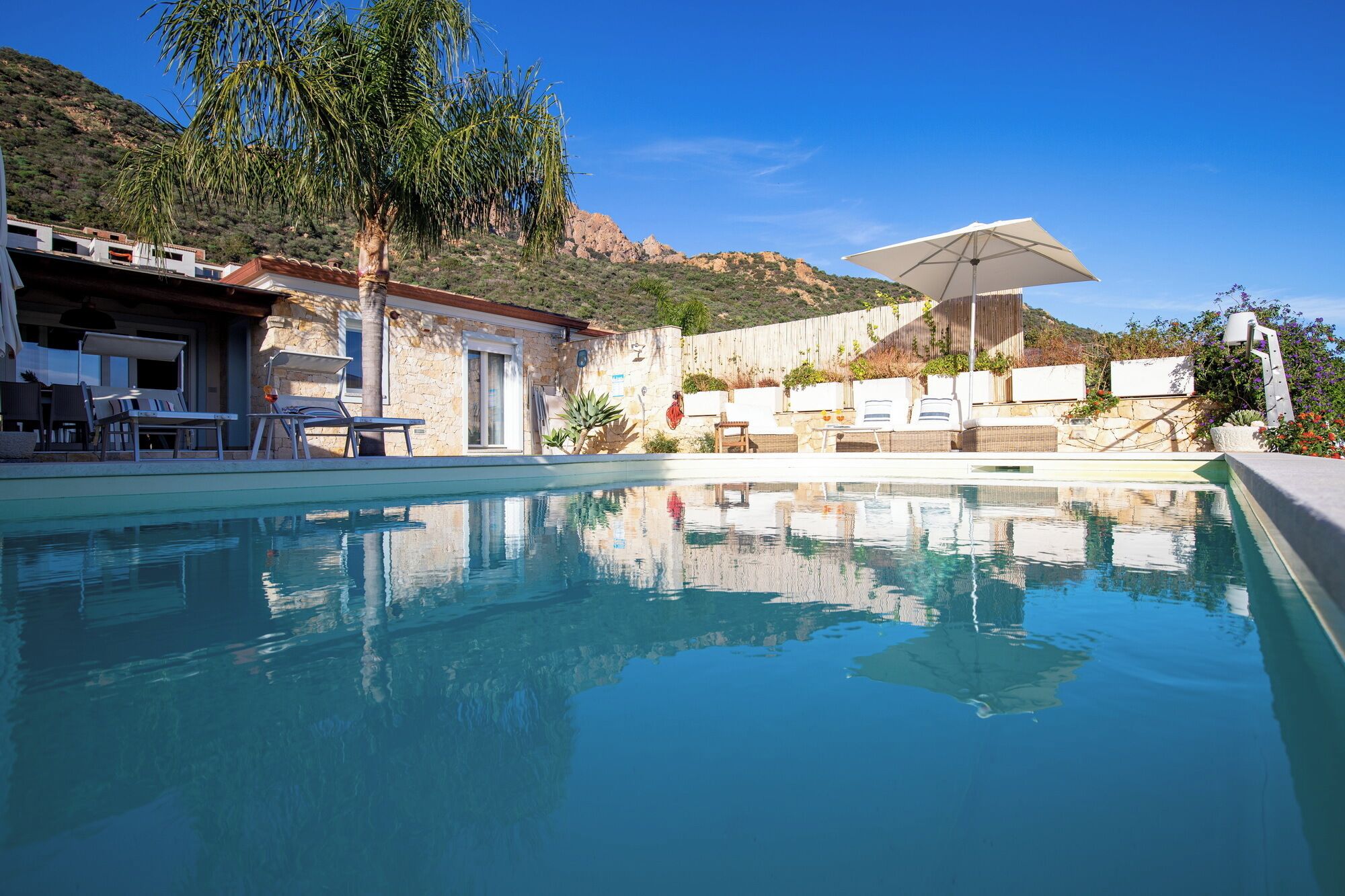 A clear swimming pool with lounge chairs and umbrellas, surrounded by a stone building and a hillside.