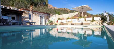 A clear swimming pool with lounge chairs and umbrellas, surrounded by a stone building and a hillside.
