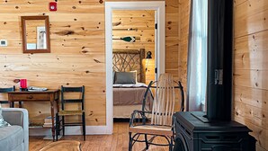 A cozy cabin interior with a wooden floor, a rustic dining table, a comfortable sofa, a wooden rocking chair, and a black wood stove. - Mrs. Otis's Cabin with Woodland View (Paul Smiths)