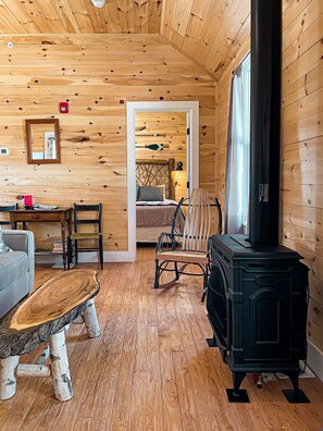 A cozy cabin interior with a wooden floor, a rustic dining table, a comfortable sofa, a wooden rocking chair, and a black wood stove.