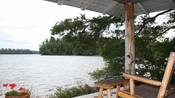 A wooden bench and a potted plant on a concrete ledge overlooking a body of water. - Mrs. Otis's Cabin with Woodland View (Paul Smiths)