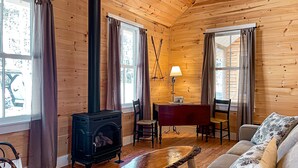 A cozy cabin with a wood-burning stove, wooden walls, a sofa, and a dining area. - Mrs. Otis's Cabin with Woodland View (Paul Smiths)