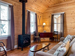 A cozy cabin with a wood-burning stove, wooden walls, a sofa, and a dining area. - Mrs. Otis's Cabin with Woodland View (Paul Smiths)