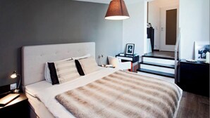 Ein modernes Schlafzimmer mit einem großen Bett, einem Nachttisch, einer Lampe, einem Schreibtisch mit Stuhl und einer Tür zu einem weiteren Raum.