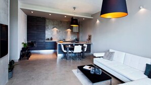 Ein modernes Wohnzimmer mit einem weißen Sofa, einem schwarzen Couchtisch und einem Essbereich, im Hintergrund befindet sich eine Küche.