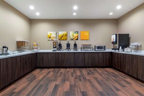 Una habitación de hotel moderna con suelo de madera, una kitchenette con cafetera, microondas y tostadora. Hay cuadros colgados en la pared y un pequeño estante con frutas.