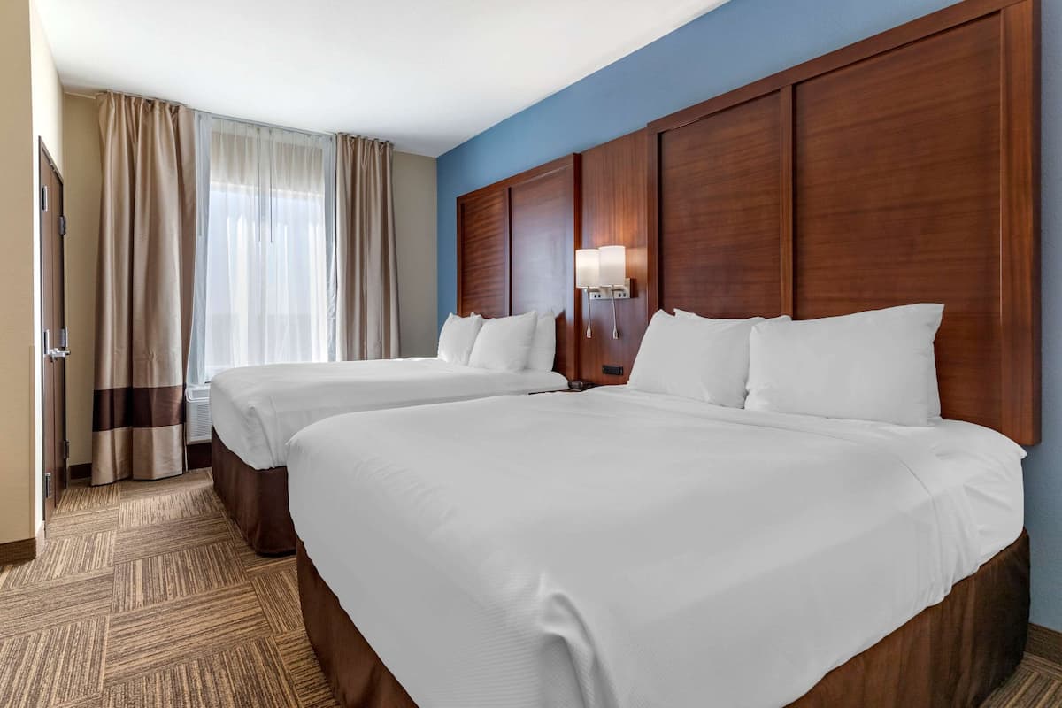 Habitación de hotel con dos camas, cabeceros de madera, un ventanal con cortinas y suelo alfombrado.