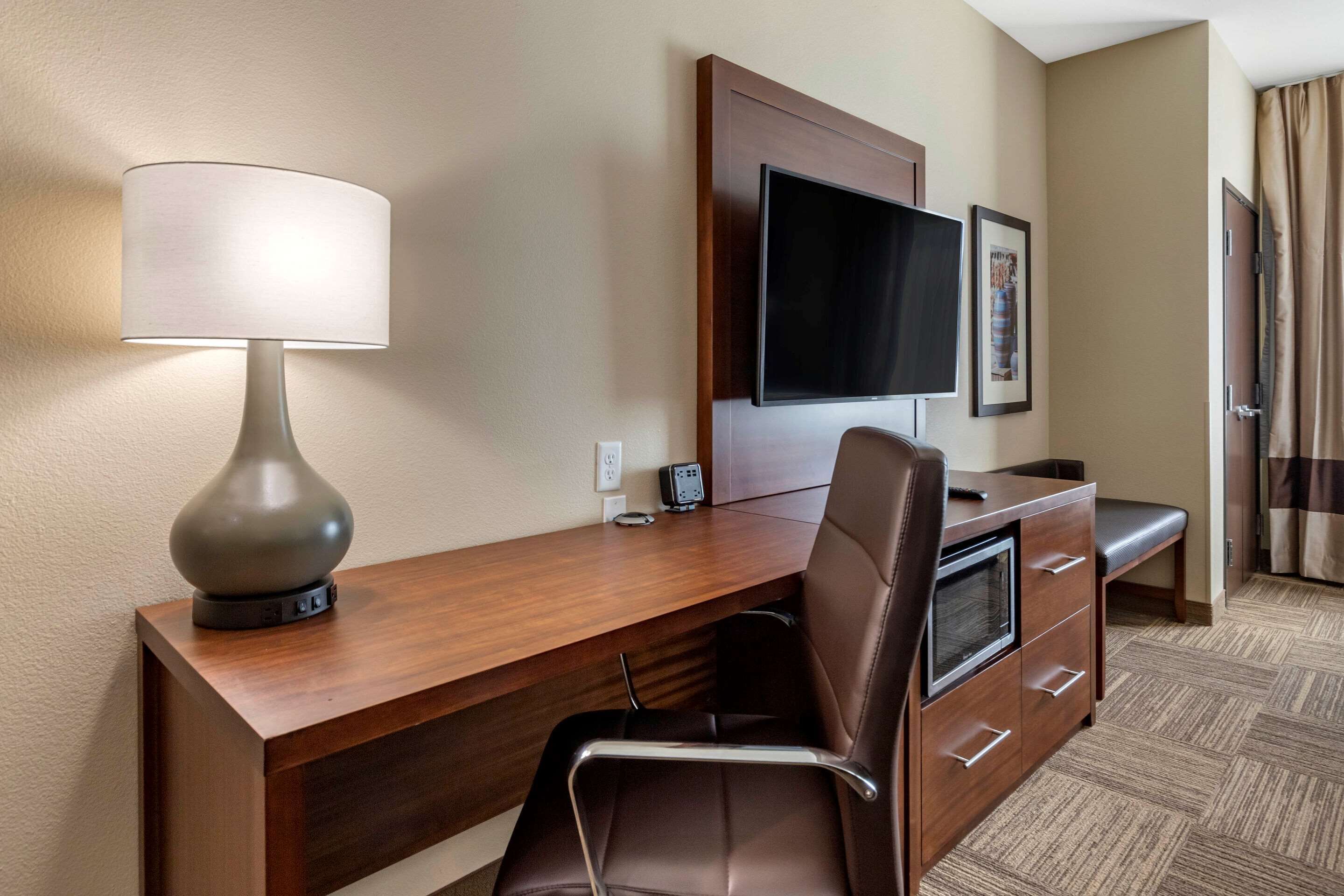 Habitación de hotel con escritorio de madera, una silla moderna, televisor de pantalla plana, lámpara y un cuadro en la pared.