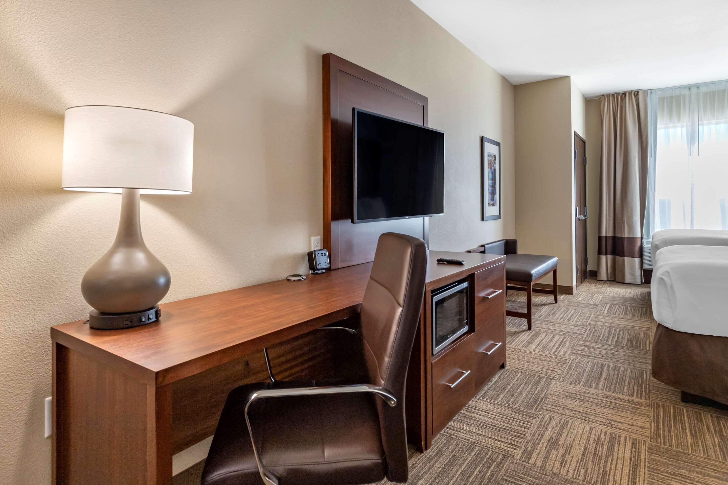 Habitación de hotel con escritorio de madera, televisor montado en la pared, cama y ventana con cortinas.