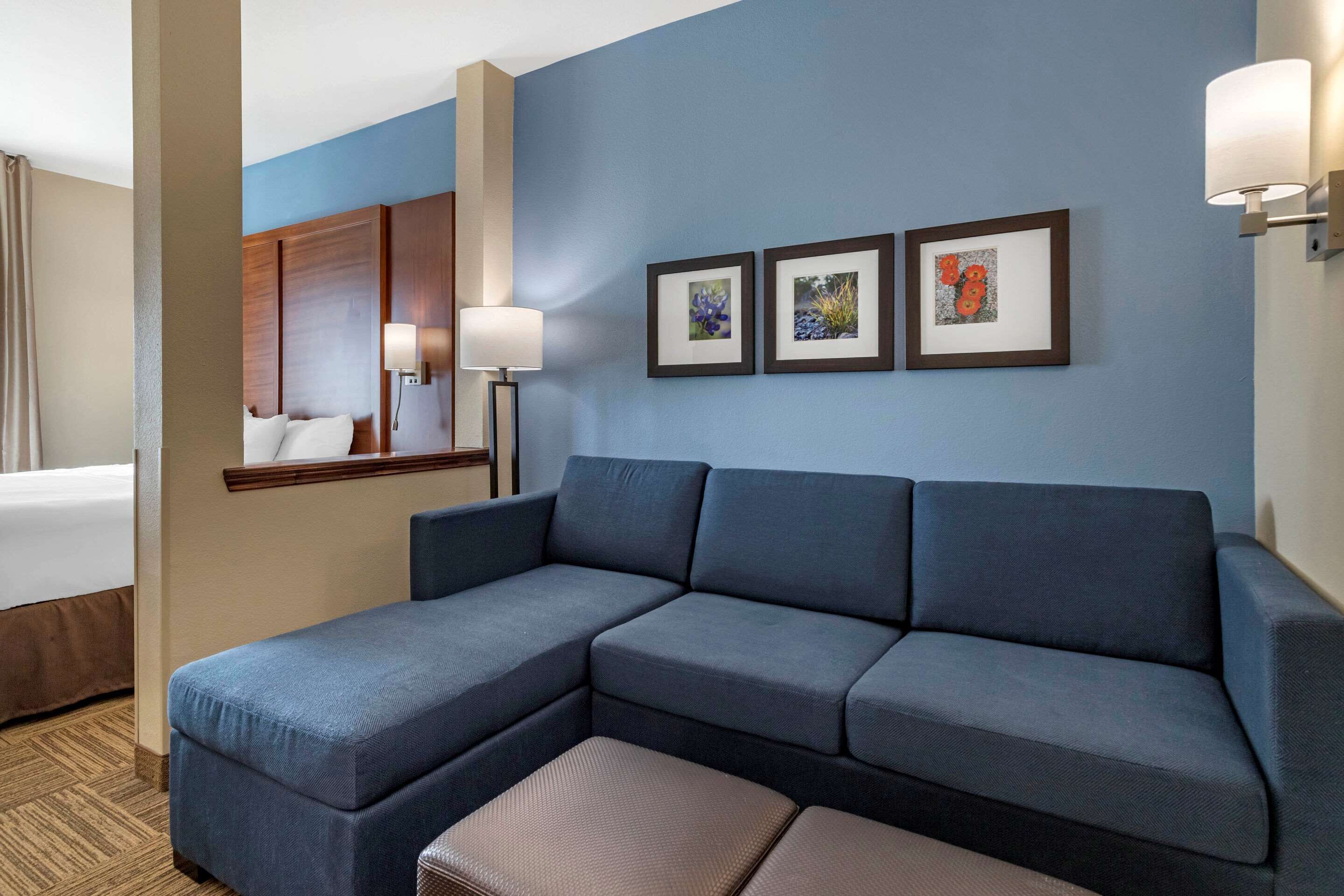 A hotel room with a large sectional sofa, a bedside table with a lamp, and two framed pictures on the wall.