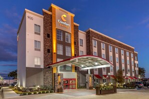 Un hotel Comfort Suites con diseño moderno, que destaca por una entrada con toldo rojo y un vestíbulo cubierto.