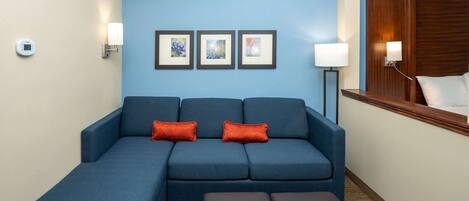 A hotel room with a blue sofa, two red cushions, a wooden headboard, and a wall-mounted thermostat.