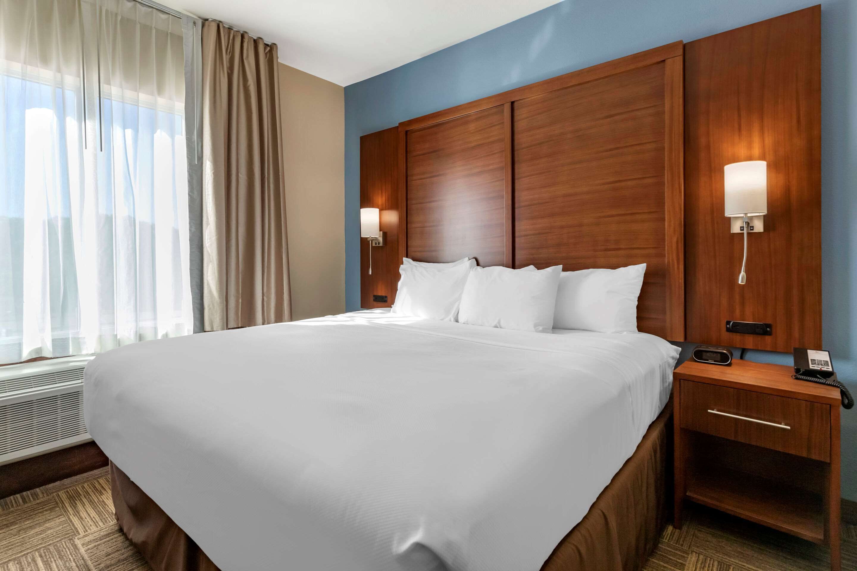 A large bed with white linens, a wooden headboard, two bedside lamps, a nightstand with a phone, and a window with sheer curtains.