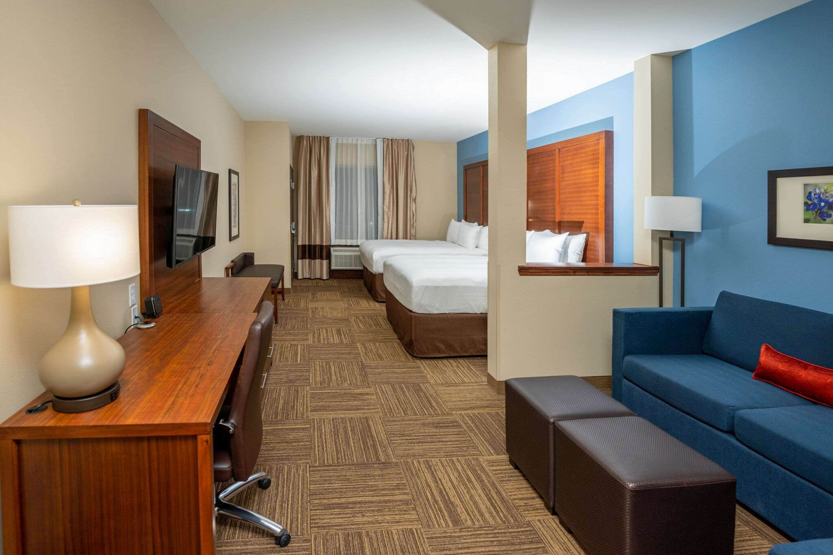 A hotel room with a wooden desk, two beds, a blue sofa, and a large window.