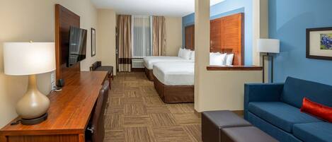 A hotel room with a wooden desk, two beds, a blue sofa, and a large window.