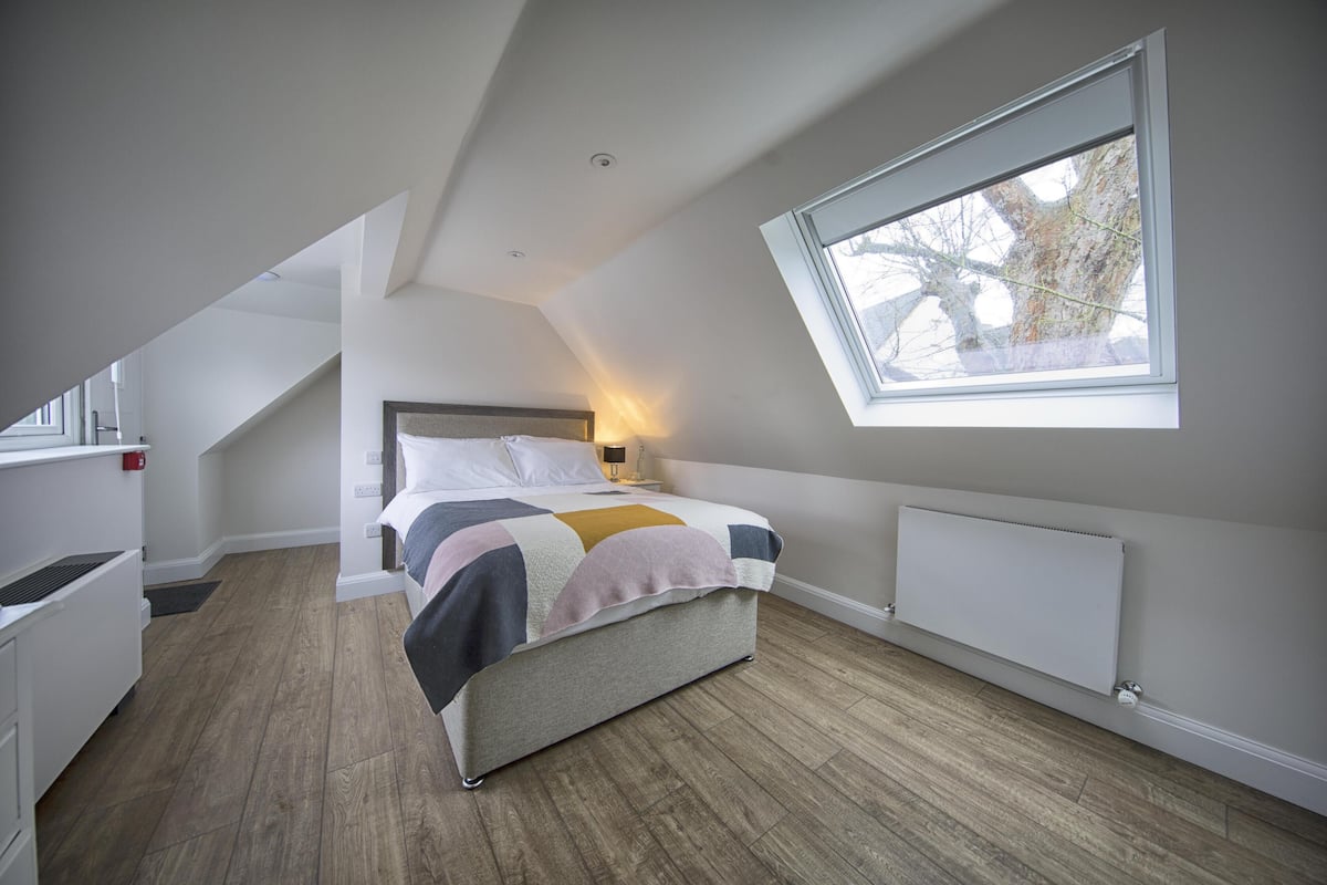 Une chambre avec un plafond en pente, une grande fenêtre, un lit recouvert d’une courtepointe, une table de chevet et un radiateur.