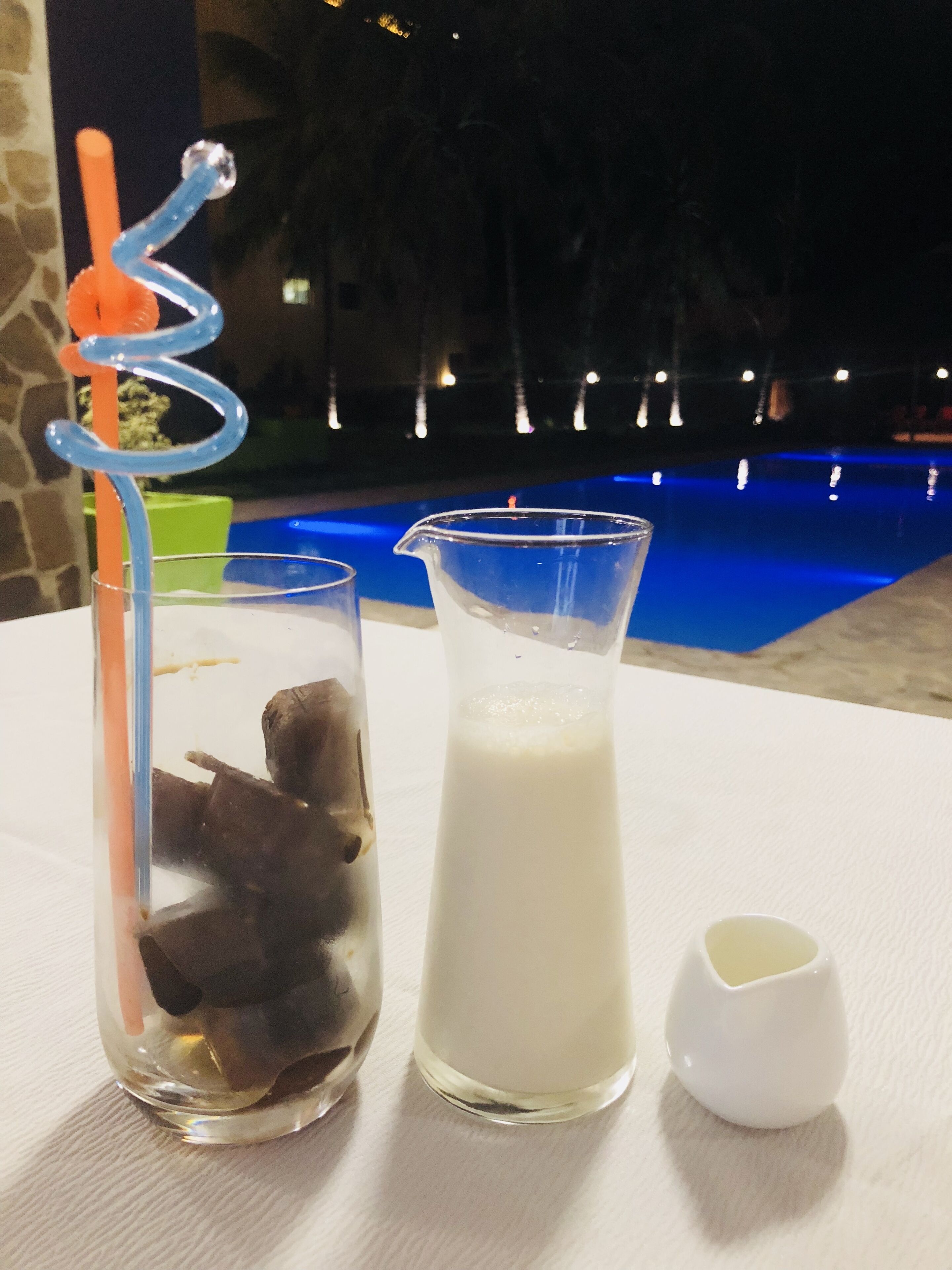 Um copo com cubos de gelo, um copo de leite e um pequeno recipiente branco sobre uma mesa perto de uma piscina.