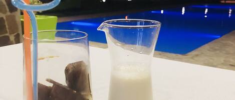 Um copo com cubos de gelo, um copo de leite e um pequeno recipiente branco sobre uma mesa perto de uma piscina.