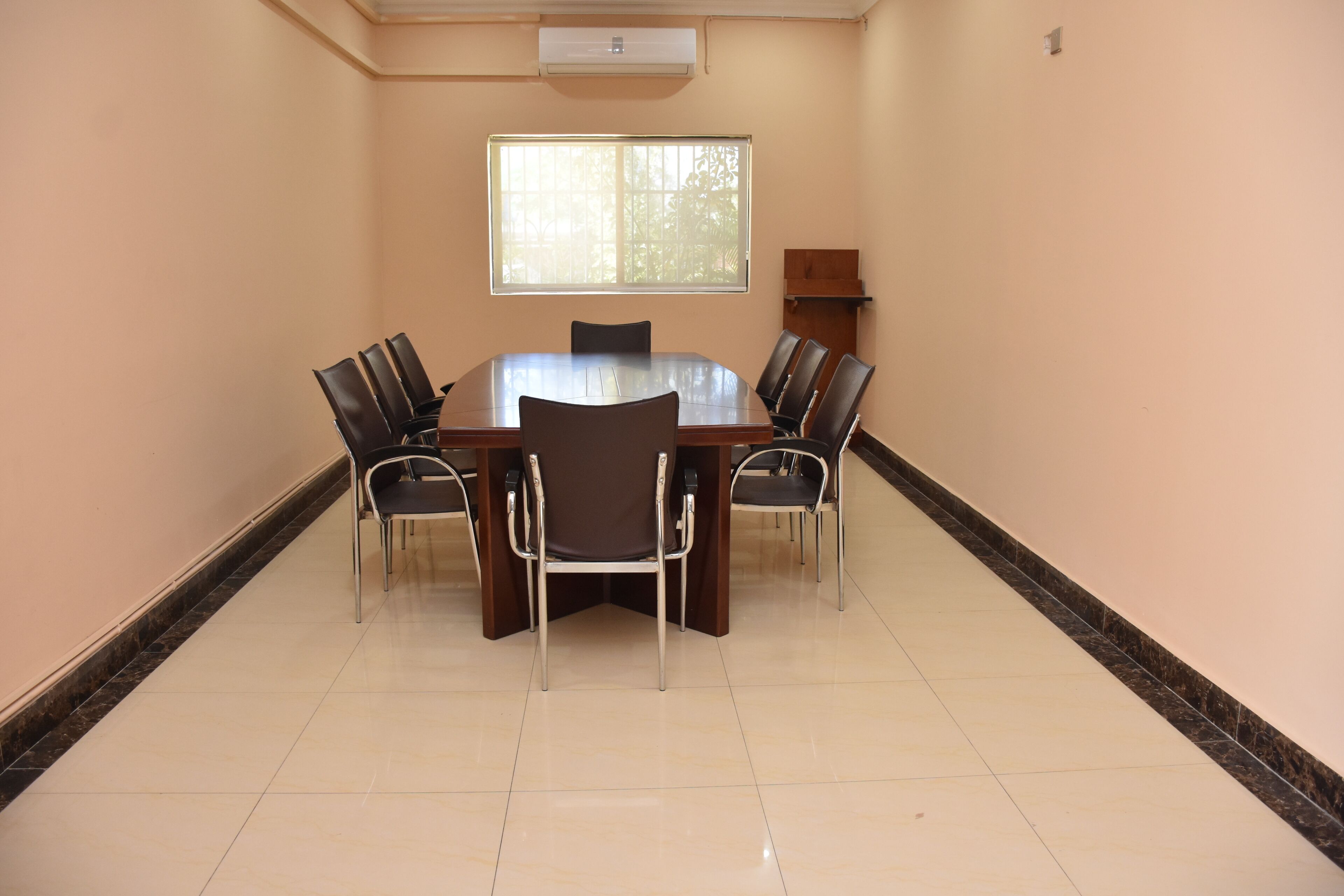 A conference room with a round table, six chairs, a window, and an air conditioning unit.