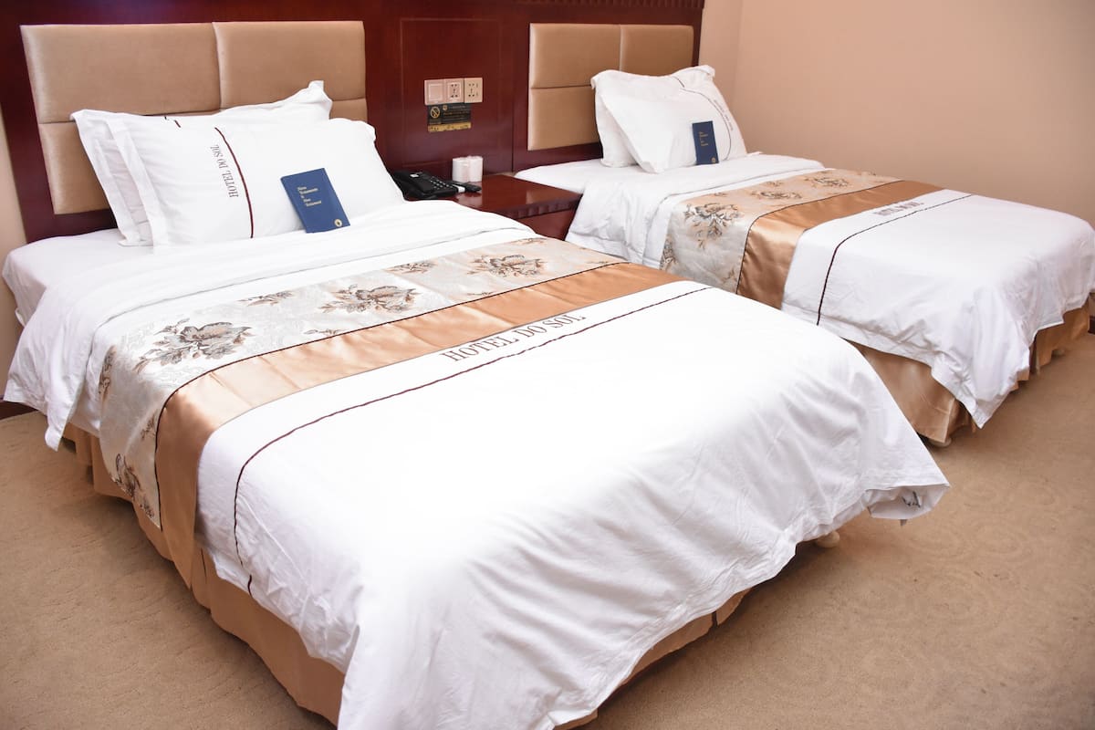Dos camas de hotel con sábanas blancas y cabeceras marrones, una mesita de noche con un teléfono y un control remoto, y una alfombra color arena.