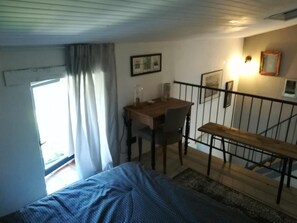 Een slaapkamer met een bed, een bureau en een stoel. Er is een raam met gordijnen, een trap en een wandlamp.