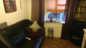 A living room with a black leather sofa, a small side table, a bookshelf, and a window with curtains. - Wexford Townhouse (Wexford)
