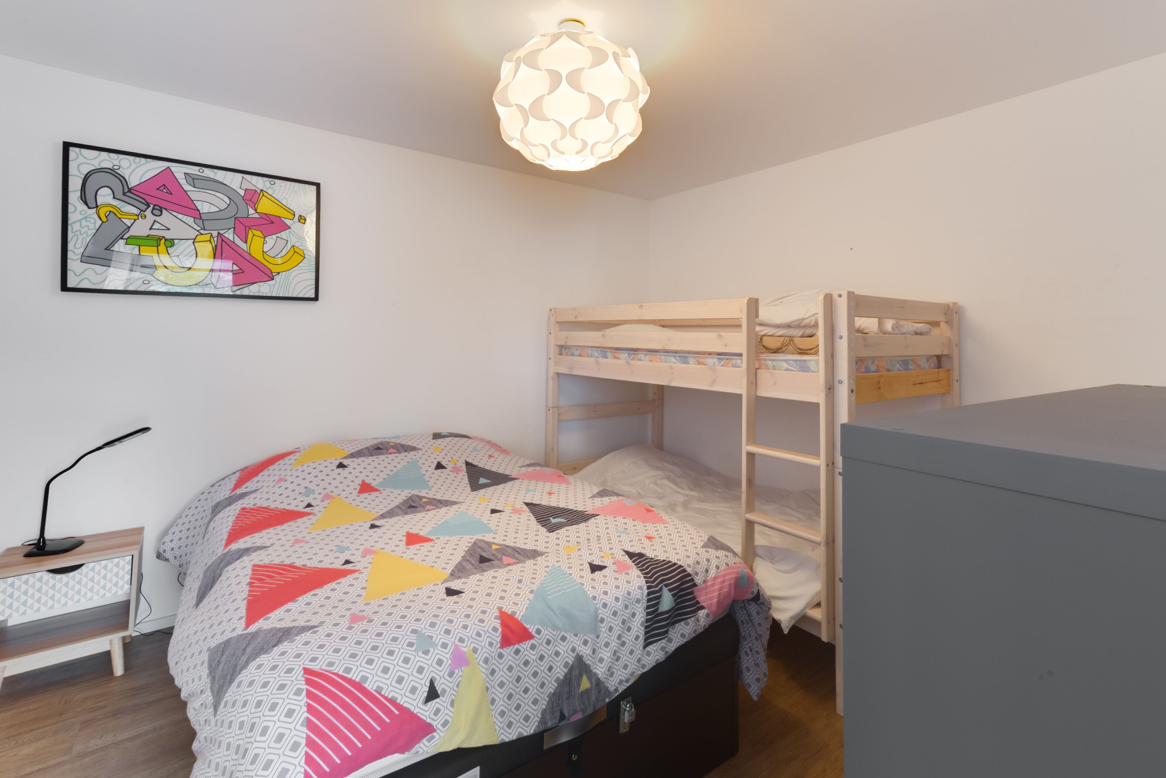 A bedroom with a bunk bed, a desk with a lamp, a wall art piece, and a hanging light fixture.