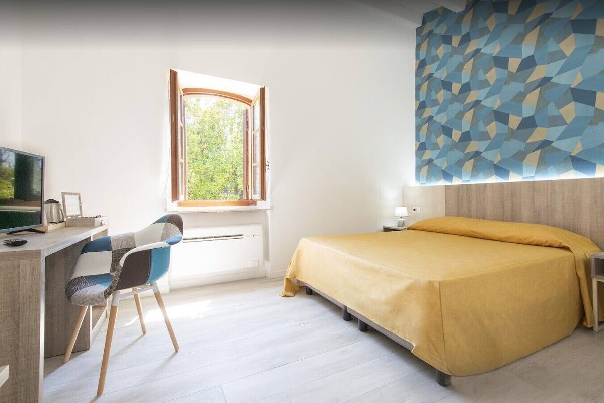 A hotel room with a bed, a desk with a chair, a TV, and a window with an open view.