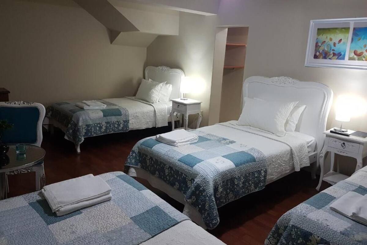 A hotel room with two beds, each with a white headboard and a blue and white quilt. There are bedside tables with lamps, a small round table with a chair, and a painting on the wall.