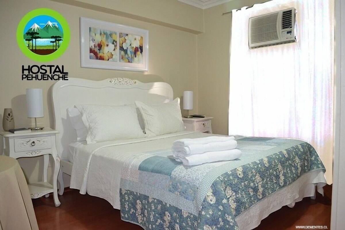 A neatly made bed with a floral bedspread, a white headboard, and two bedside tables with lamps. A framed abstract painting hangs above the bed, and an air conditioning unit is mounted on the wall.