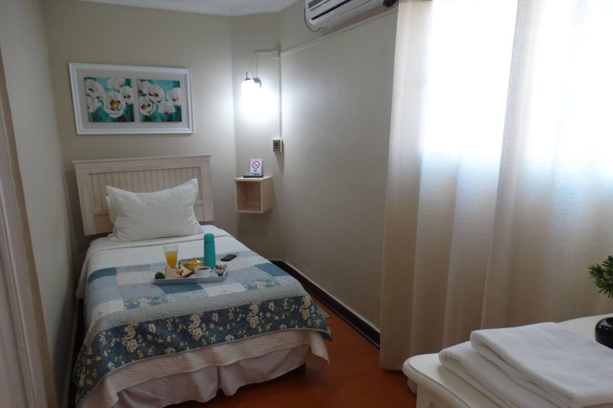 A hotel room with a bed, a bedside table with a phone, a painting on the wall, and a window with curtains.