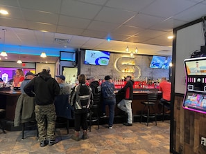 A bar area with multiple screens, a counter, and a variety of bottles.