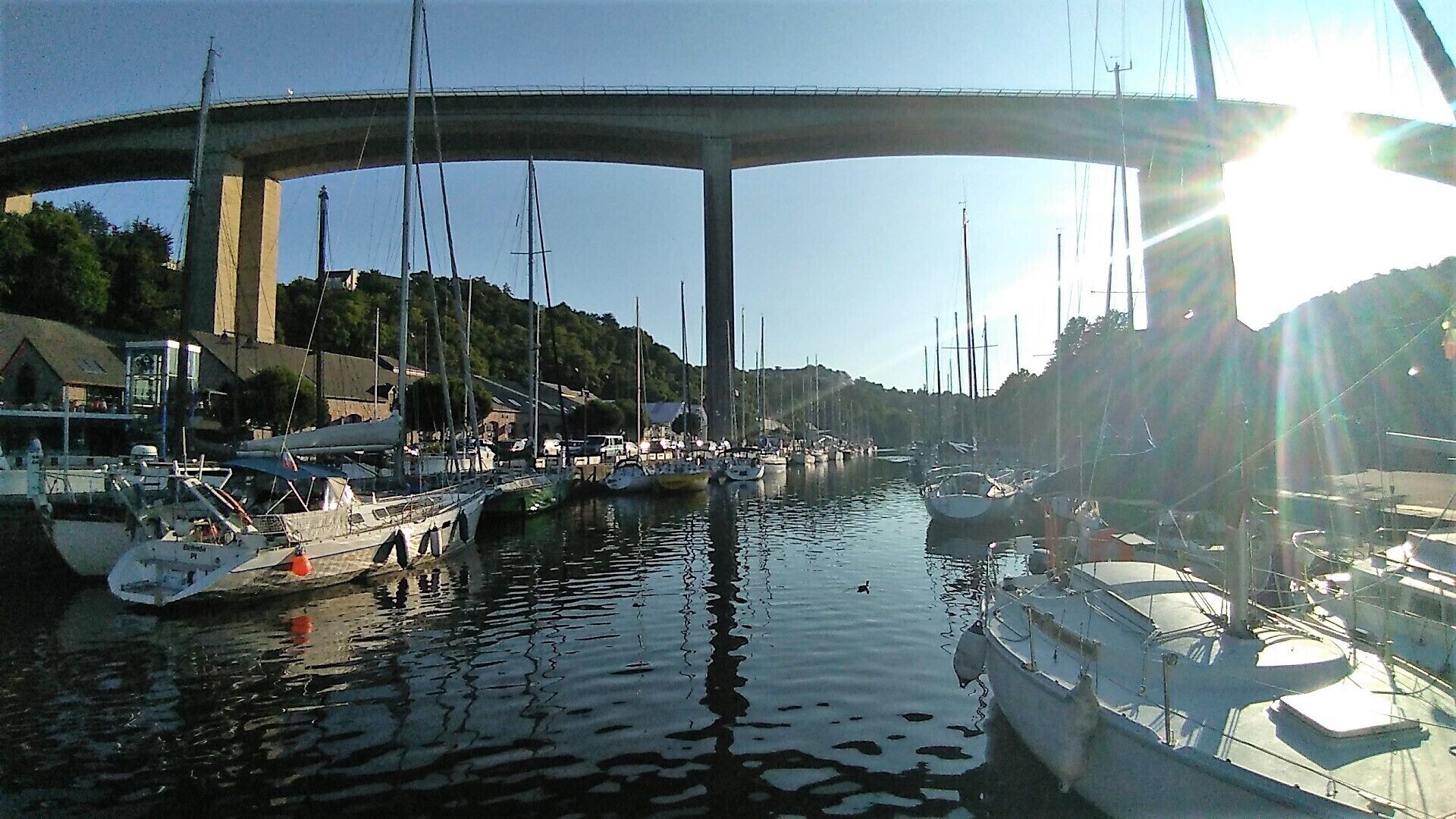 A marina with boats docked under a bridge, calm water, and a clear sky.