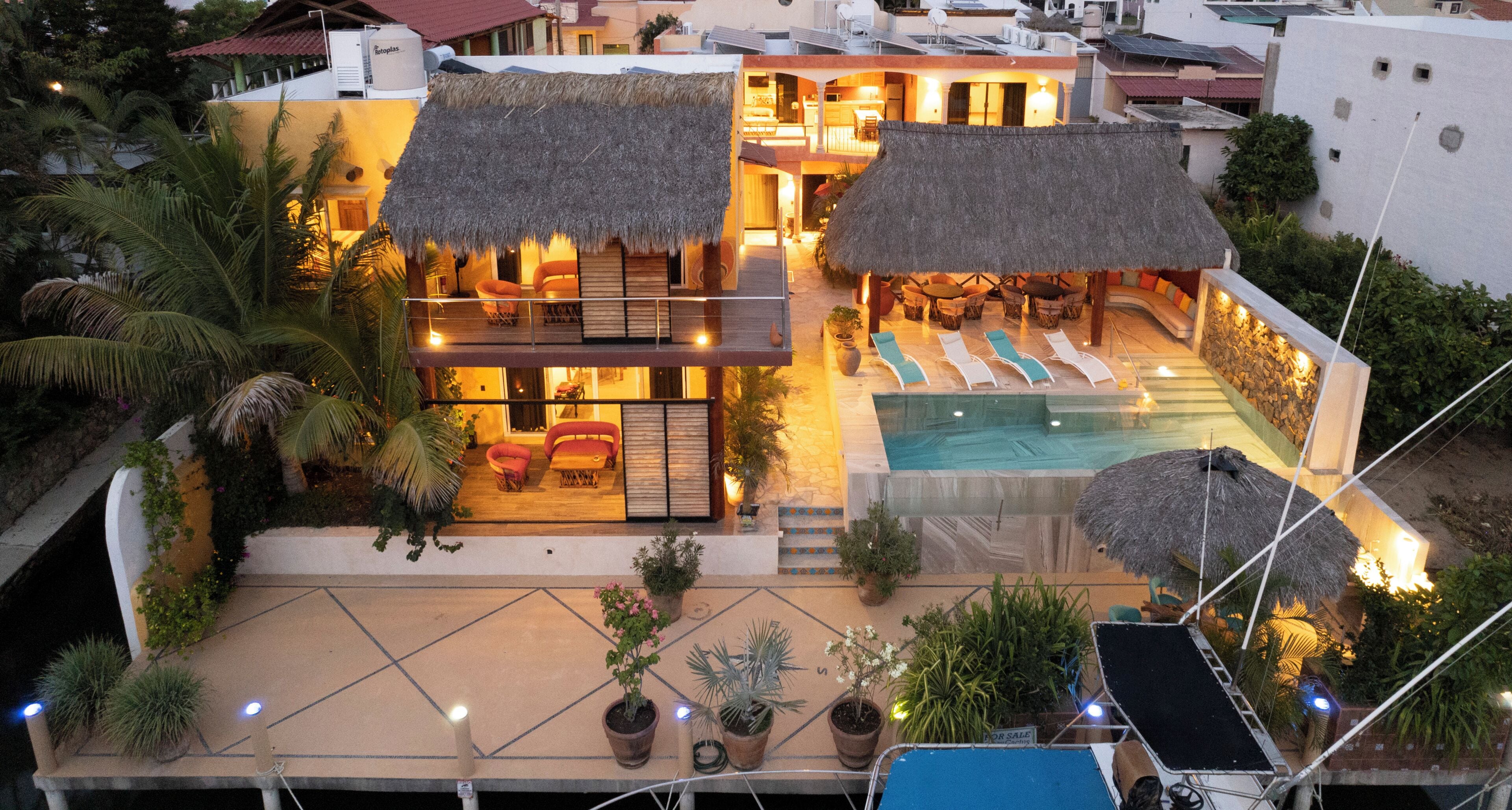 Une maison à deux étages avec une piscine, des constructions à toits de chaume et un espace salon extérieur.