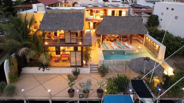Ein zweistöckiges Haus mit Pool, strohgedeckten Gebäuden und Sitzgelegenheiten im Freien.