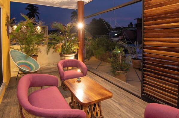 An outdoor seating area with two cushioned chairs and a wooden table, surrounded by potted plants and a view of the cityscape.