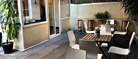 Un espacio de patio al aire libre con mesas de madera y sillas blancas, rodeado de plantas en macetas y una escalera que lleva a un nivel superior.
