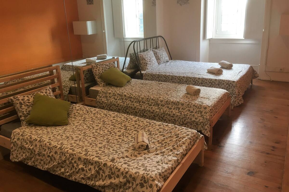 Habitación de hotel con tres camas individuales, cada una con colcha y almohadas estampadas. Hay un banco de madera y una pequeña mesa con lámpara. El piso es de madera y hay una ventana con cortinas.