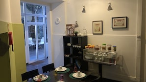 Un coin repas avec une table dressée pour quatre personnes, une bibliothèque noire, un présentoir en verre contenant des produits alimentaires et une fenêtre donnant sur un espace extérieur aménagé avec des sièges.