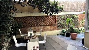 Un espace extérieur aménagé en terrasse, avec des tables en bois et des chaises blanches, entouré de plantes en pot et d’un mur en briques.