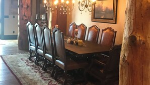 Een eetruimte met een grote houten tafel, zes stoelen, een kroonluchter en een schilderij aan de muur.