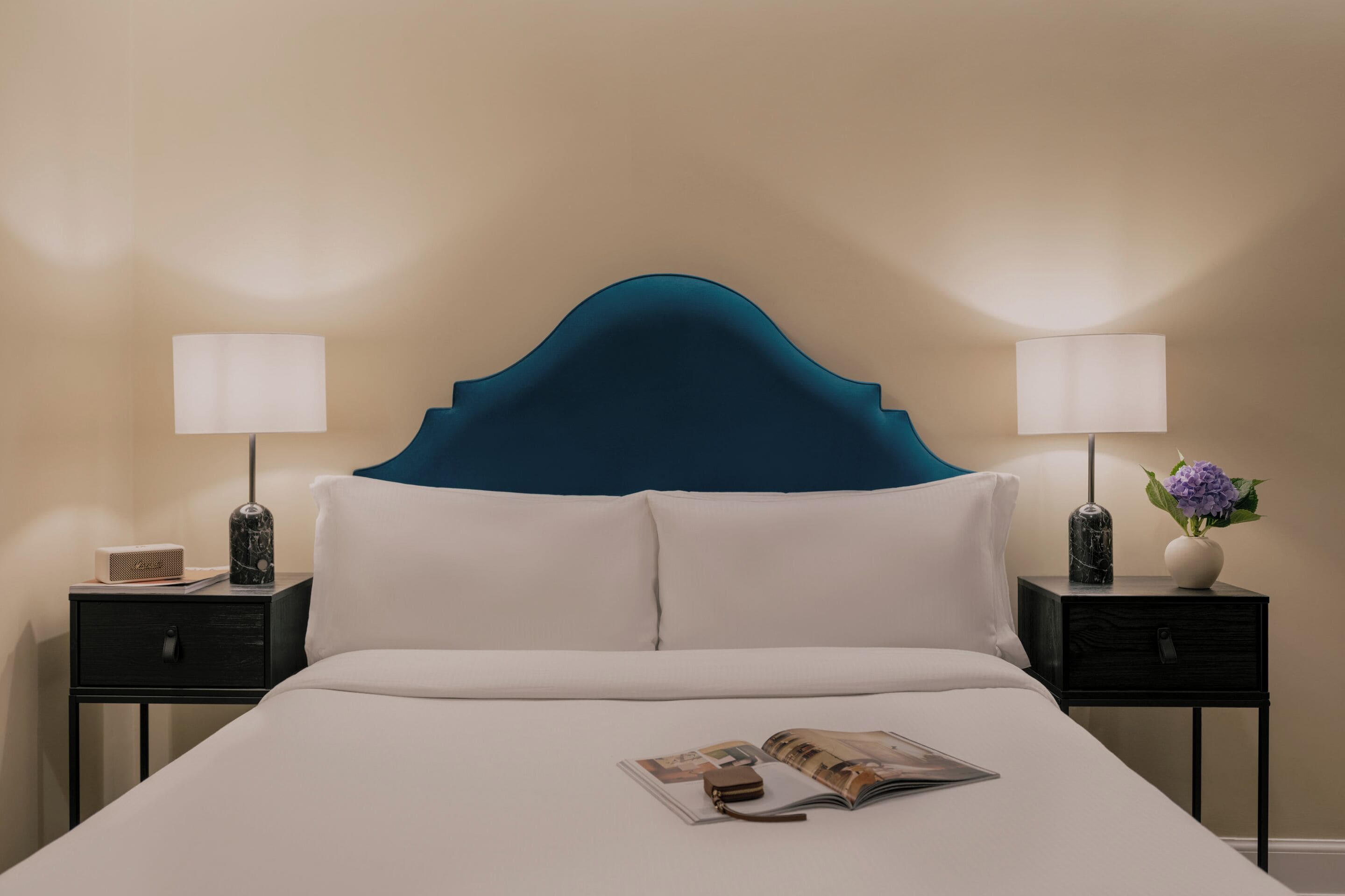 A neatly made bed with a teal headboard, two bedside tables with lamps, a small vase with a purple flower, and a magazine on the bed.