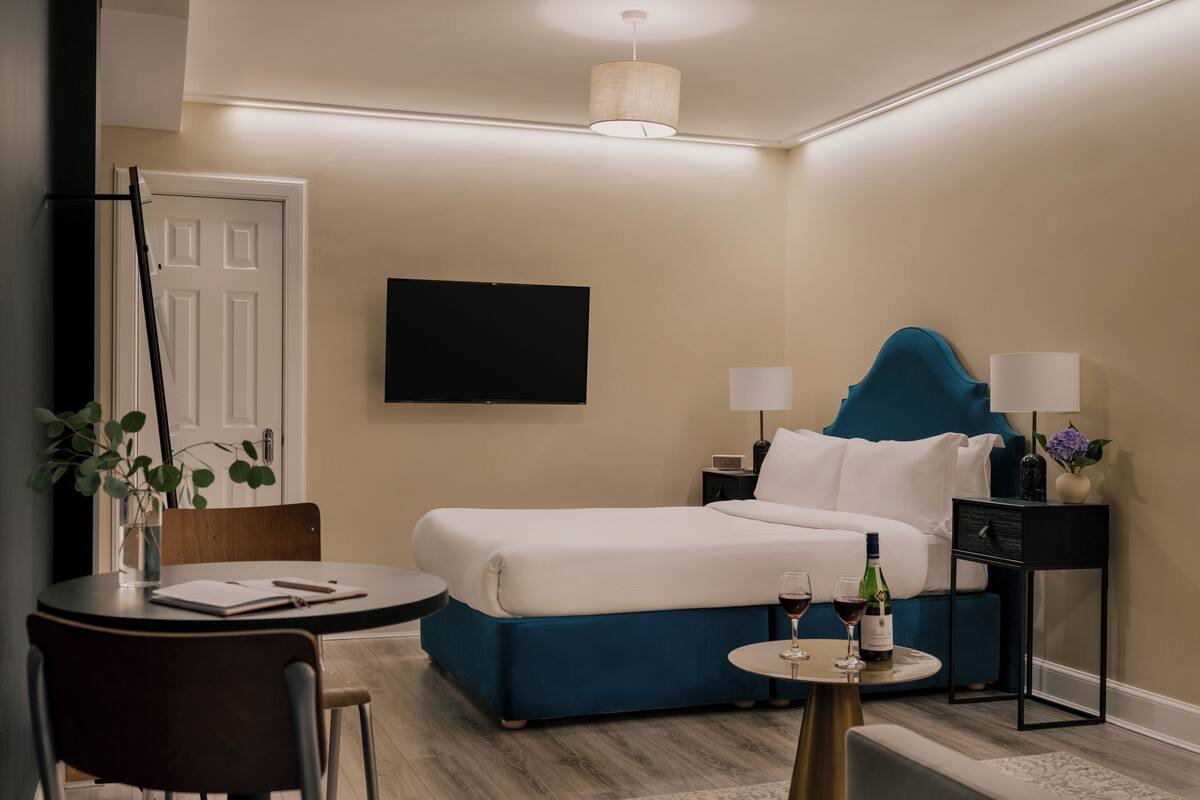 Habitación de hotel con cama, mesitas de noche, televisor, escritorio con silla y una pequeña mesa con una botella y vasos.